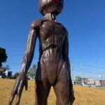 a imagem mostra um et de escultura. atrás, o cenário de uma grama amarelada, o céu azul e carros e estabelecimentos