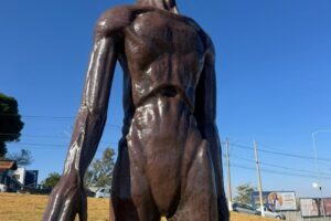 a imagem mostra um et de escultura. atrás, o cenário de uma grama amarelada, o céu azul e carros e estabelecimentos