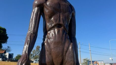 a imagem mostra um et de escultura. atrás, o cenário de uma grama amarelada, o céu azul e carros e estabelecimentos