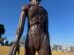 a imagem mostra um et de escultura. atrás, o cenário de uma grama amarelada, o céu azul e carros e estabelecimentos