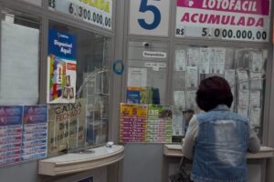 a imagem mostra uma mulher em um a lotérica. De fundo, a banca com anúncios divulgando os prêmios.