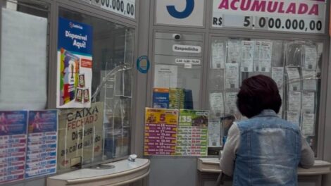 a imagem mostra uma mulher em um a lotérica. De fundo, a banca com anúncios divulgando os prêmios.