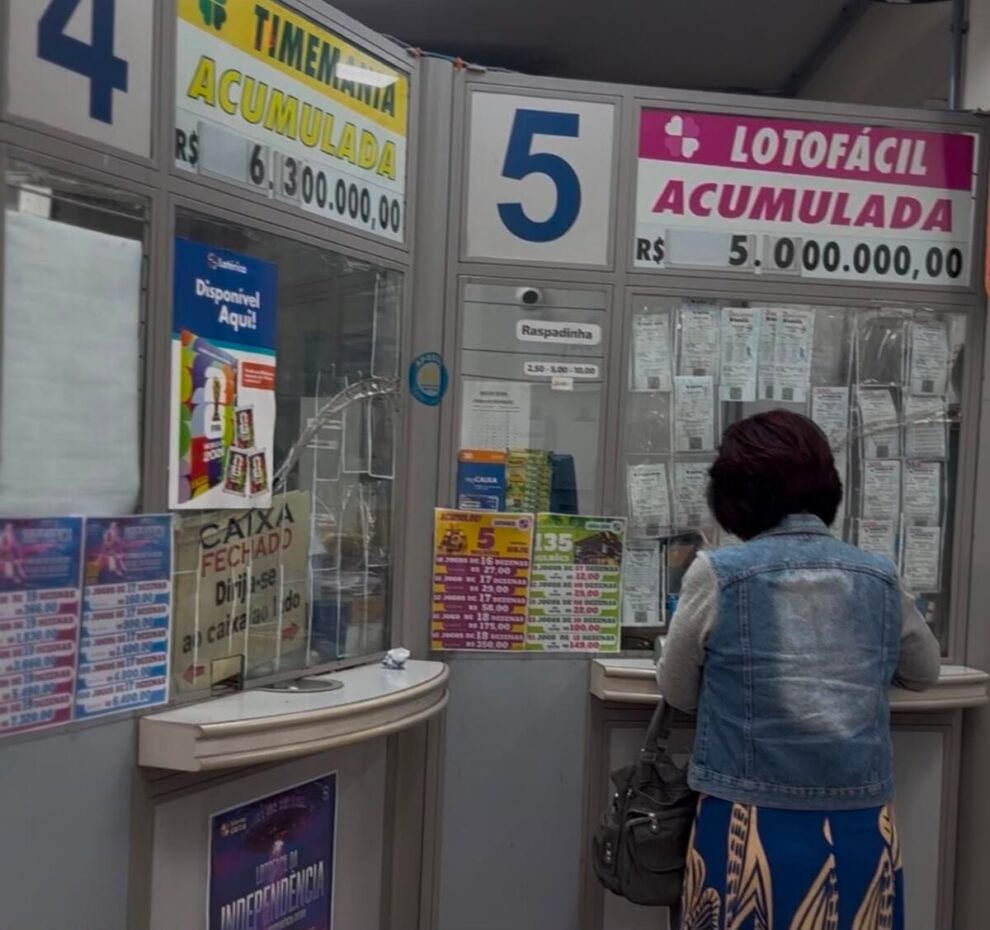 a imagem mostra uma mulher em um a lotérica. De fundo, a banca com anúncios divulgando os prêmios.